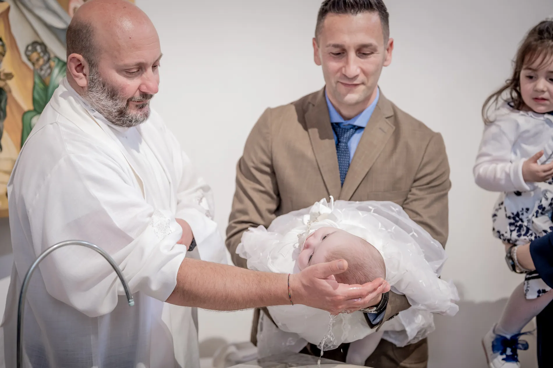 Durante il rito il sacerdote versa l’acqua battesimale sul bambino, sostenuto da un uomo in giacca