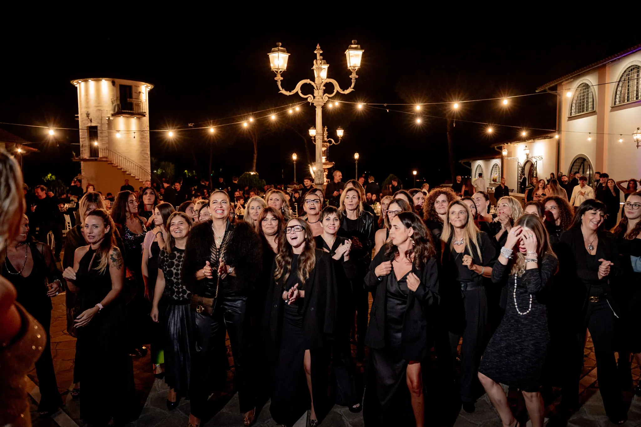 Grande folla di invitati sotto i filari di luci in un cortile durante un evento serale