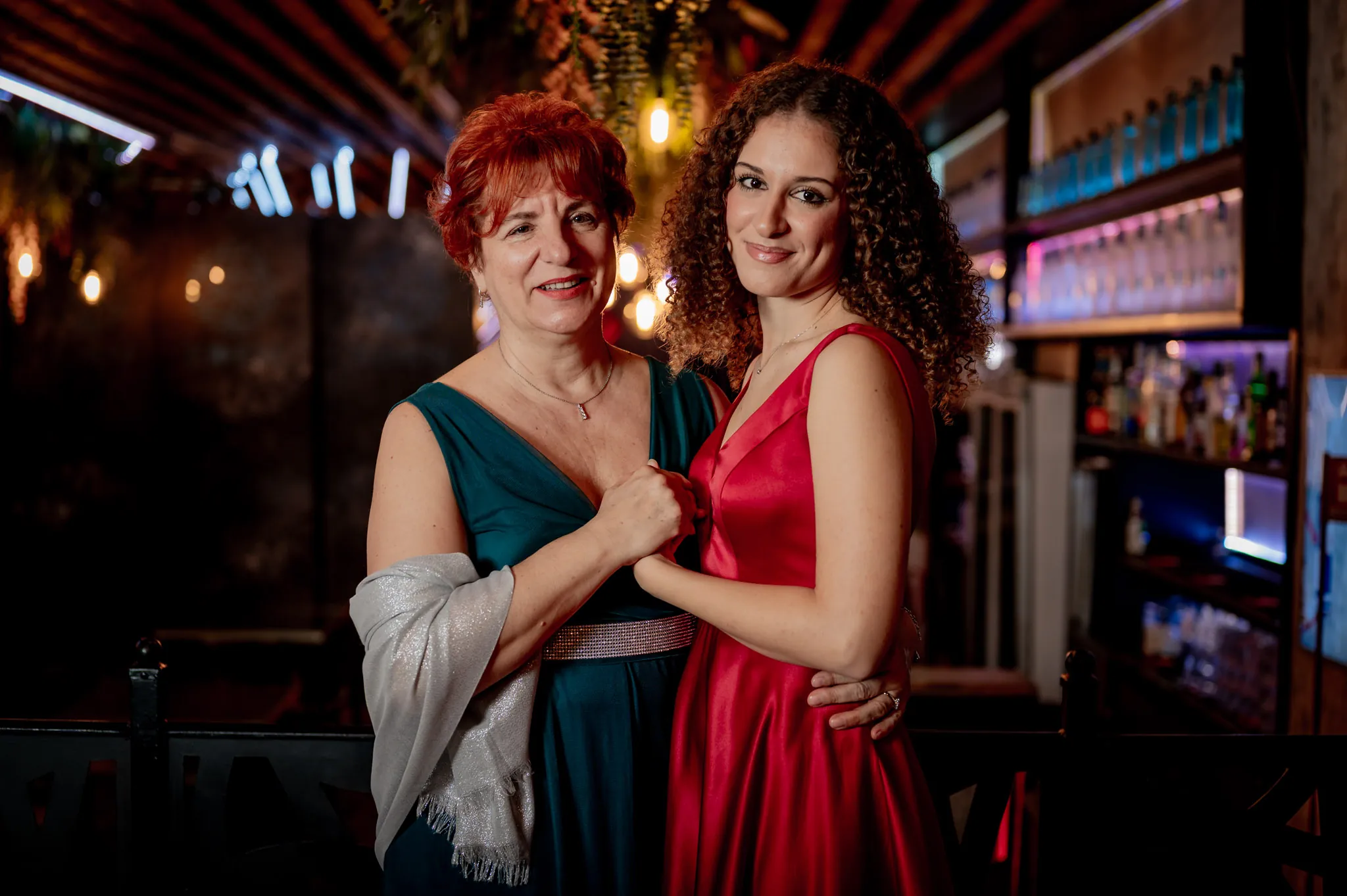 Due donne in abito lungo sorridono vicine accanto al bancone del bar