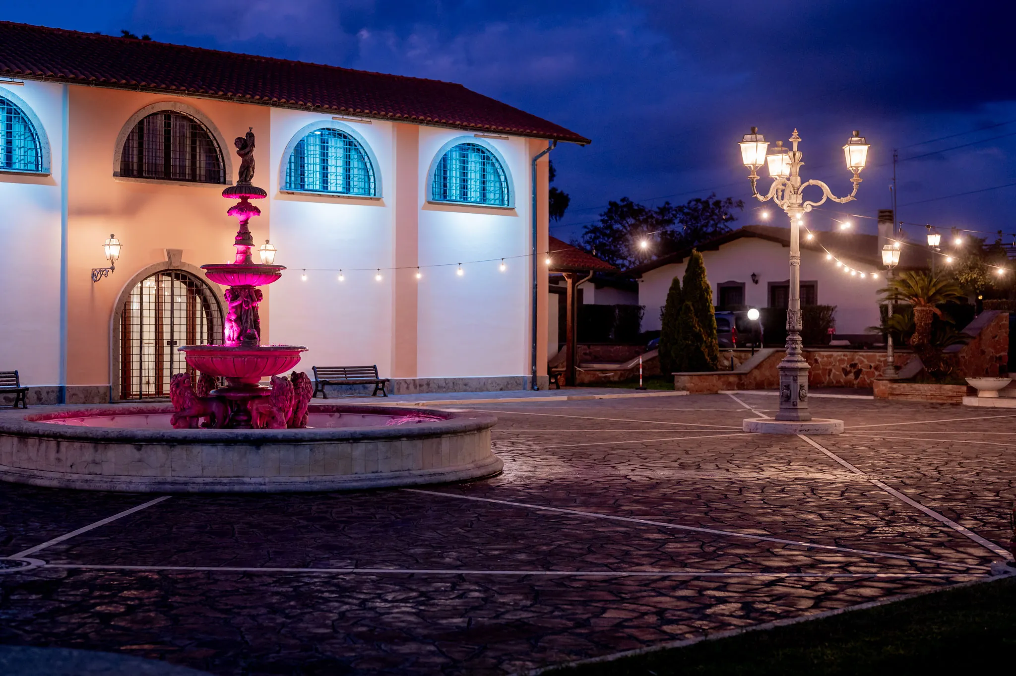 Cortile di una villa con fontana illuminata di rosa e lampioni all’ora blu