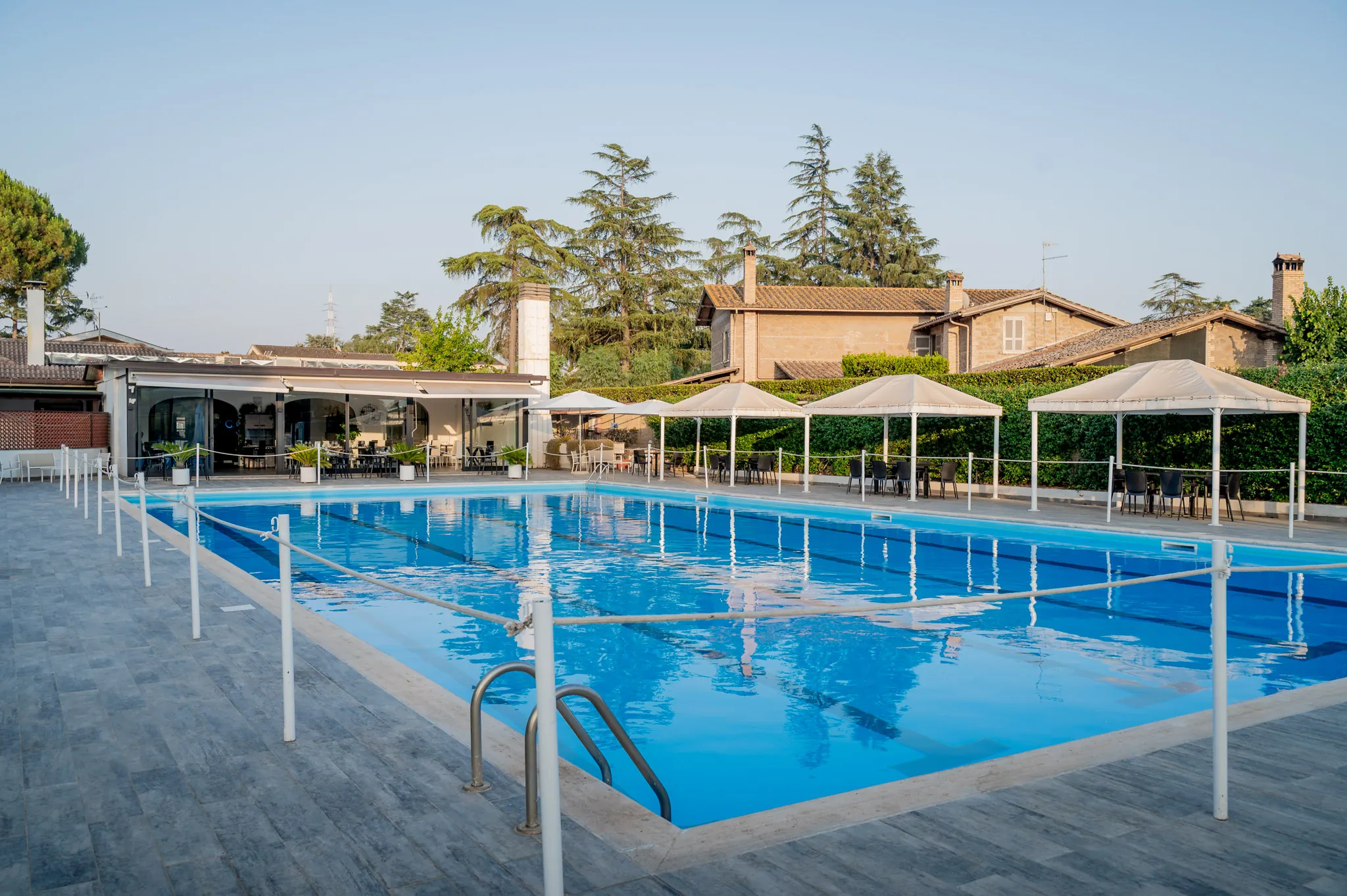 Piscina di una villa con ombrelloni bianchi e alberi sullo sfondo, nel pomeriggio