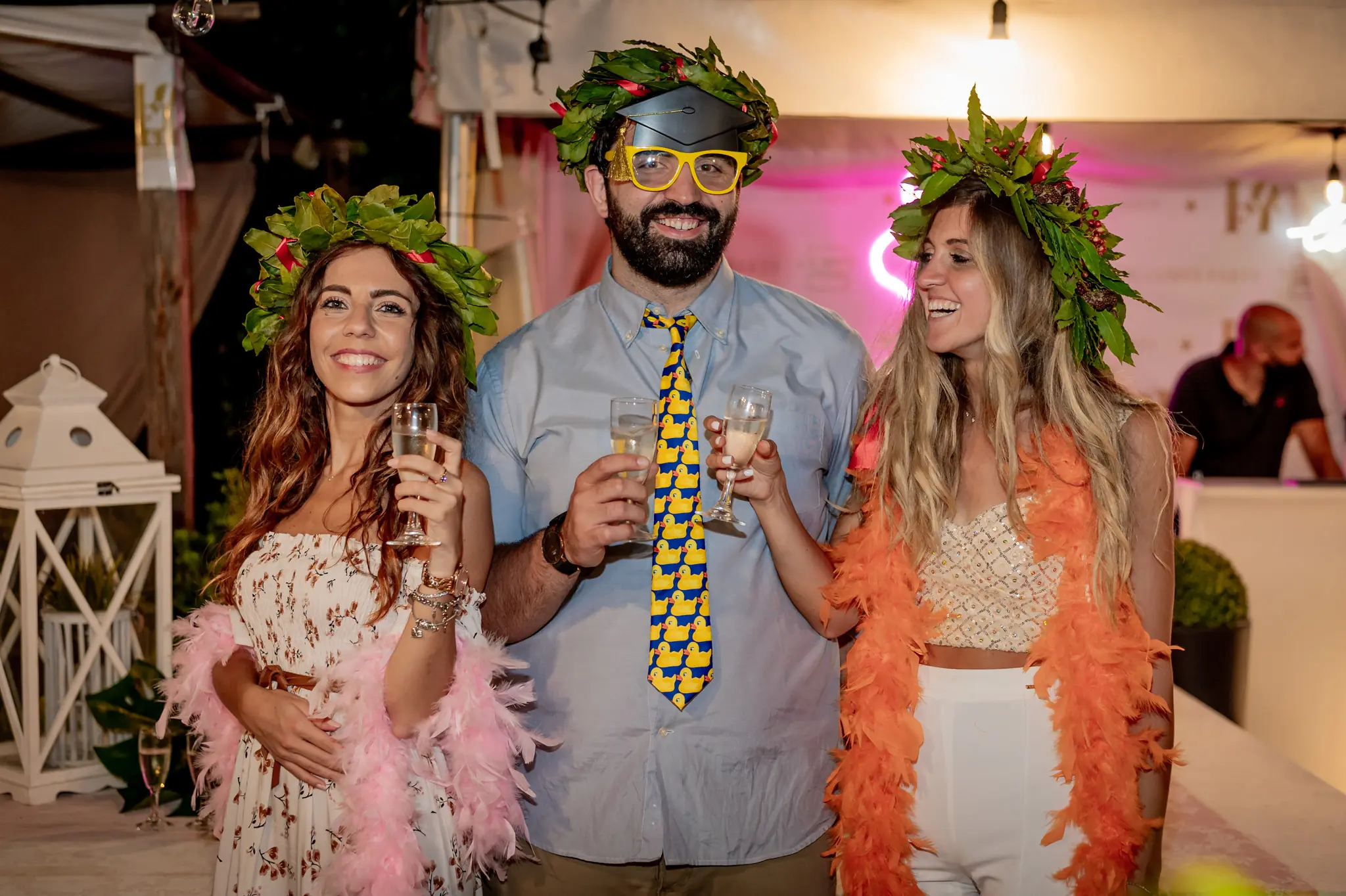 Due amiche con la corona d’alloro e un amico con occhiali e cravatta buffi brindano alla festa